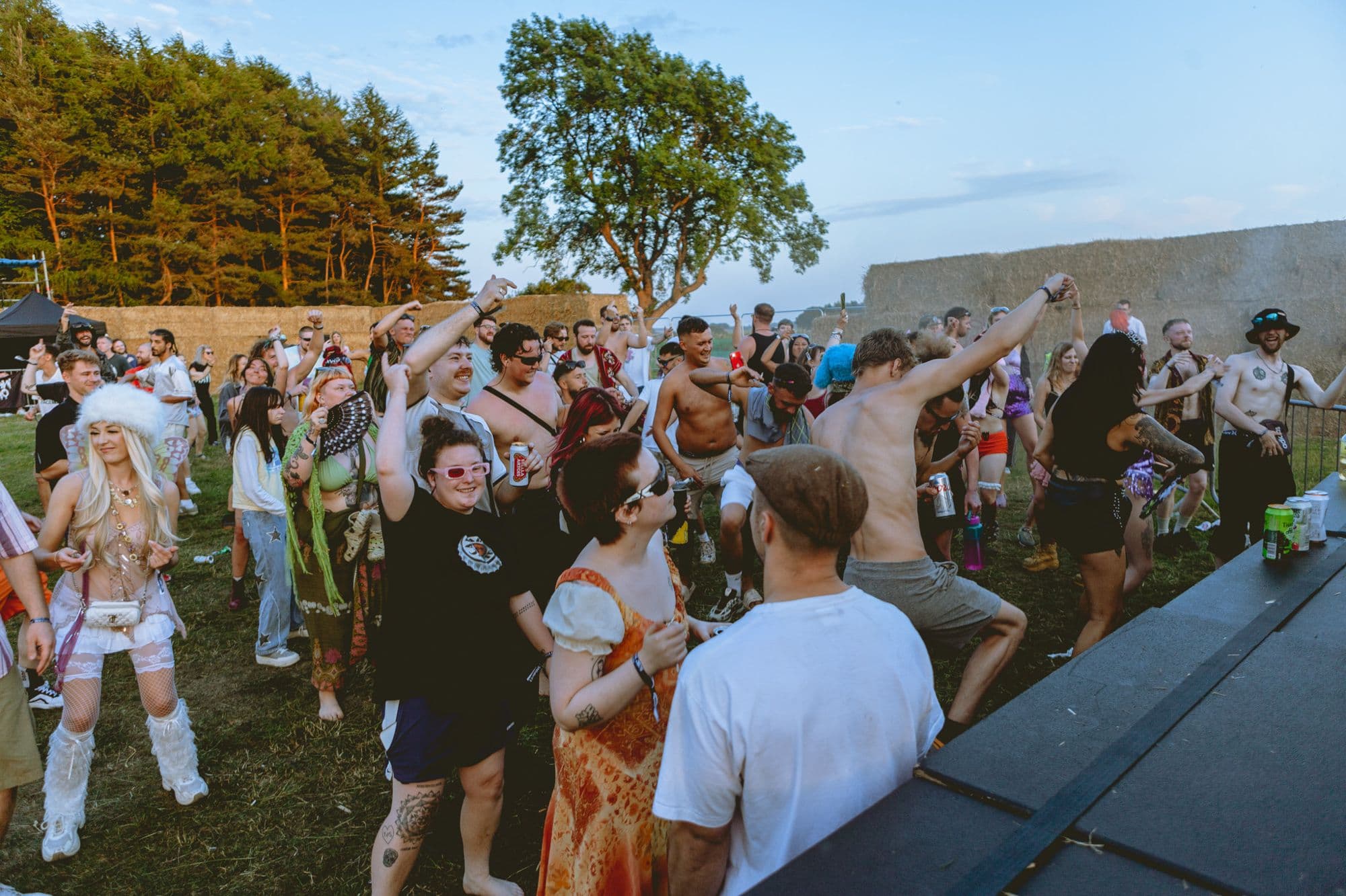 Crowd dancing in a field in front of tall pine trees and hay bales